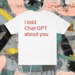 Puede incluir: Camiseta blanca con el texto rojo "I told Chat GPT about you". La camiseta se muestra sobre un fondo de graffiti colorido. La camiseta es una prenda informal.