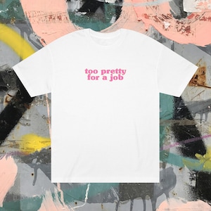 Puede incluir: Camiseta blanca con la frase rosa "too pretty for a job". La camiseta tiene cuello redondo y está hecha de una tela suave. El fondo es una colorida pared de graffiti.
