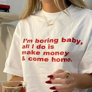 Puede incluir: Camiseta blanca con texto rojo que dice "Soy aburrido bebé, todo lo que hago es ganar dinero y volver a casa."