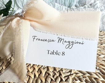 May include: A white card with the text "Francesca Maggioni Table 8" on it. The card is sitting on a woven wicker placemat and is decorated with a peach-coloured ribbon with frayed edges.