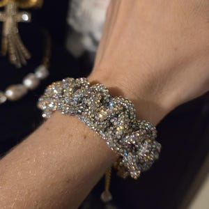 May include: A bracelet featuring a thick, braided design completely covered in small, iridescent, light-reflecting crystals. The piece has a dense, textured appearance with a shimmering, multi-colored finish that catches the light from various angles.