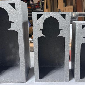 May include: Three gray, rectangular decorative boxes of varying sizes. Each box features a cutout in the shape of an arched silhouette with triangular accents at the top. The boxes have a textured, speckled appearance.