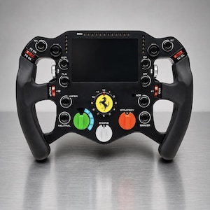 Può includere: Un volante da corsa di Formula 1 nero con un display digitale centrale, un logo Ferrari giallo e vari pulsanti e selettori rotativi etichettati in rosso, verde, arancione e bianco. Il dispositivo include etichette come MGU, TC FIX, TC OL, WIPER, NEUTRAL, STRATEGY, MARKER ed ENGINE.