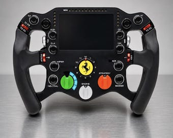May include: A black Formula 1 racing steering wheel featuring a central digital display, a yellow Ferrari logo, and various labeled buttons and rotary switches in red, green, orange, and white. The device includes text labels like MGU, TC FIX, TC OL, WIPER, NEUTRAL, STRATEGY, MARKER, and ENGINE.