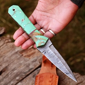 May include: A green and brown patterned handle dagger with a Damascus steel blade. The blade is double-edged and has a pointed tip.