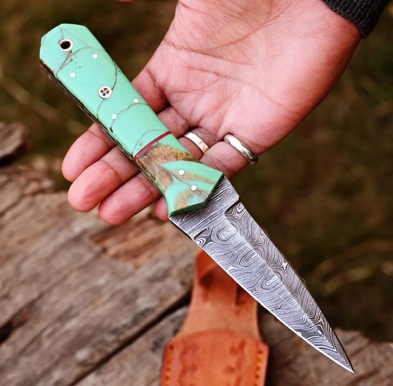 May include: A green and brown patterned handle dagger with a Damascus steel blade. The blade is double-edged and has a pointed tip.