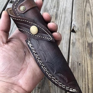 May include: A brown leather knife sheath with white stitching and a decorative pattern. The sheath has a brass button closure.
