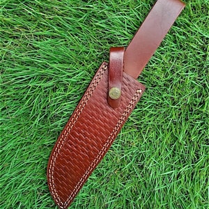 May include: A brown leather knife sheath with a stitched design and a belt loop. The sheath has a brass button closure.