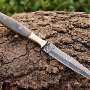 May include: A black and grey handle dagger with a brass bolster and a damascus steel blade. The blade has a pointed tip and a slightly curved edge. The dagger is lying on a piece of rough bark.