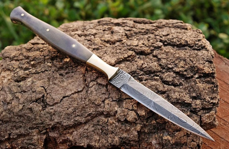 May include: A black and grey handle dagger with a brass bolster and a damascus steel blade. The blade has a pointed tip and a slightly curved edge. The dagger is lying on a piece of rough bark.
