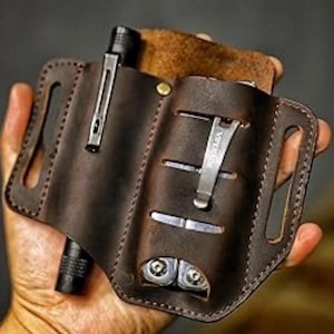 May include: A brown leather multi-tool holder with a belt loop. The holder has two slots for tools, one for a pen, and one for a flashlight. The holder is made of high-quality leather and is designed to be durable and long-lasting.