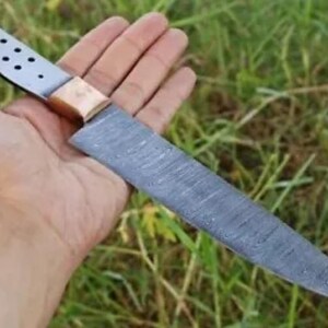 May include: A long, silver knife with a copper handle and a patterned blade. The knife is held in a hand against a green grassy background.