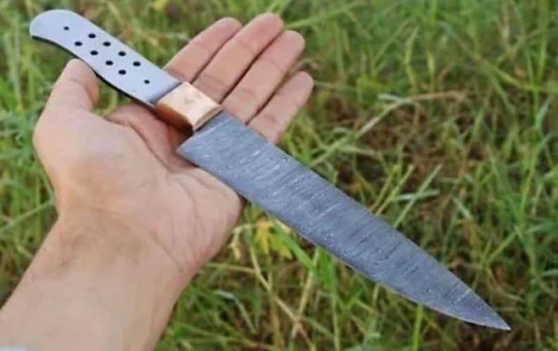 May include: A long, silver knife with a copper handle and a patterned blade. The knife is held in a hand against a green grassy background.