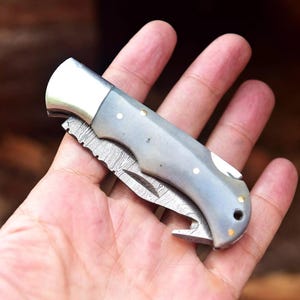 May include: A gray and silver folding knife with a decorative blade pattern. The knife has a curved handle with brass rivets and a silver bolster. The blade is partially open, revealing the intricate design. The knife is held in a hand.