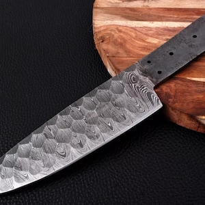 May include: A close-up of a Damascus steel knife blank with a hammered finish. The blade is approximately 9 inches long and has a rounded tip. The blank is resting on a wooden cutting board.