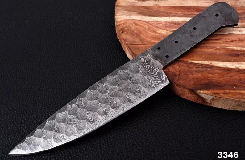 May include: A close-up of a Damascus steel knife blank with a hammered finish. The blade is approximately 9 inches long and has a rounded tip. The blank is resting on a wooden cutting board.