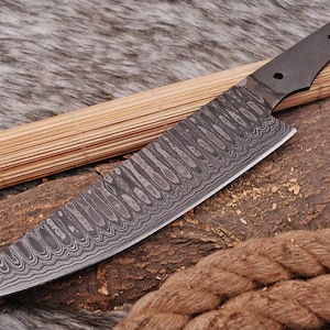 May include: A long, black-handled kitchen knife with a Damascus steel blade. The blade has a wavy pattern and is very sharp. The knife is resting on a wooden surface.