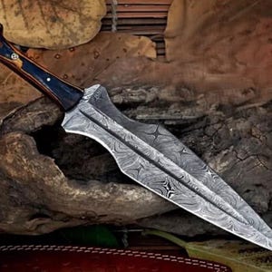 May include: A Damascus steel dagger with a dark wood handle. The blade has a distinctive pattern and a pointed tip. The handle is made of wood with a dark stain.