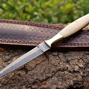 May include: A  dagger with a white bone handle and a  damascus steel blade. The dagger is resting on a piece of wood and is in a brown leather sheath. The number 1320 is visible in the top left corner of the image.