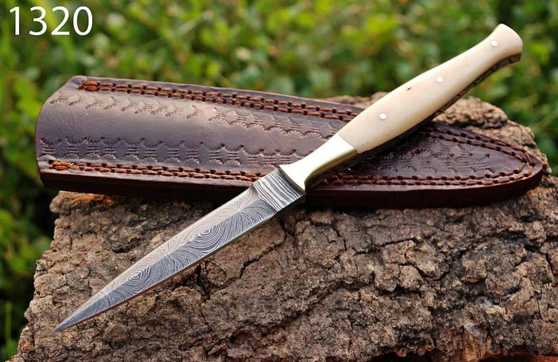 May include: A  dagger with a white bone handle and a  damascus steel blade. The dagger is resting on a piece of wood and is in a brown leather sheath. The number 1320 is visible in the top left corner of the image.