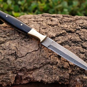 May include: A black and brass handled dagger with a Damascus steel blade. The blade has a wavy pattern and is sharp. The dagger is resting on a piece of tree bark.