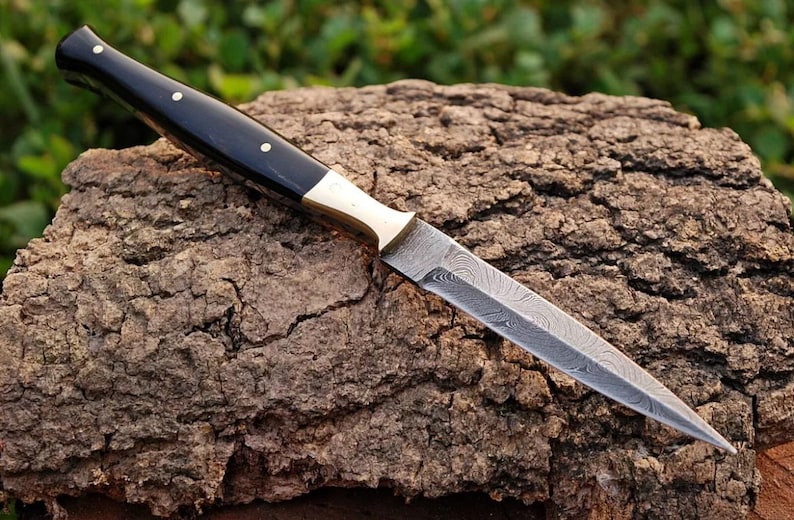 May include: A black and brass handled dagger with a Damascus steel blade. The blade has a wavy pattern and is sharp. The dagger is resting on a piece of tree bark.