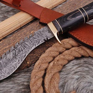 Op de afbeelding: Een Damascus staal mes met een zwart houten handvat en een leren schede. Het mes heeft een gekarteld lemmet en een gouden metalen accent.