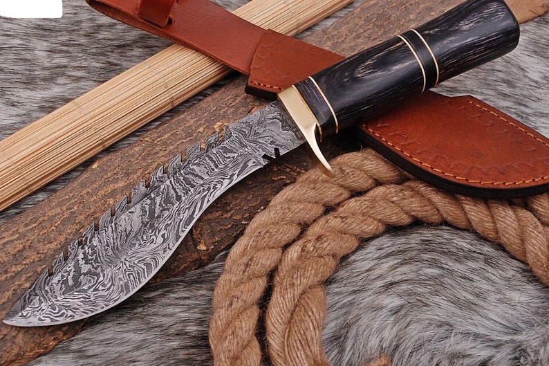 Op de afbeelding: Een Damascus staal mes met een zwart houten handvat en een leren schede. Het mes heeft een gekarteld lemmet en een gouden metalen accent.