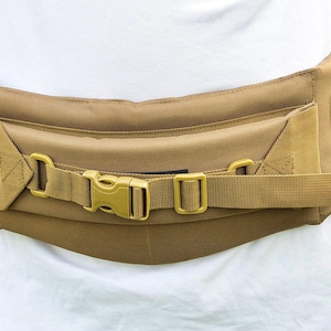 May include: Tan tactical belt with a padded design. Features a secure buckle closure and adjustable straps. The belt is designed to be worn around the waist and has multiple attachment points for accessories.