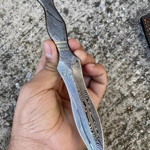 May include: A Damascus steel knife with a wavy pattern. The handle is dark with a curved design, and the blade has a pointed tip. The knife is held in a hand against a gray background.