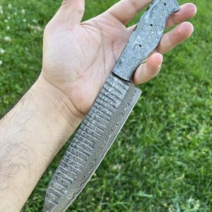 May include: A close-up of a silver knife with a patterned blade and a textured handle. The knife is held in a hand against a green grassy background.