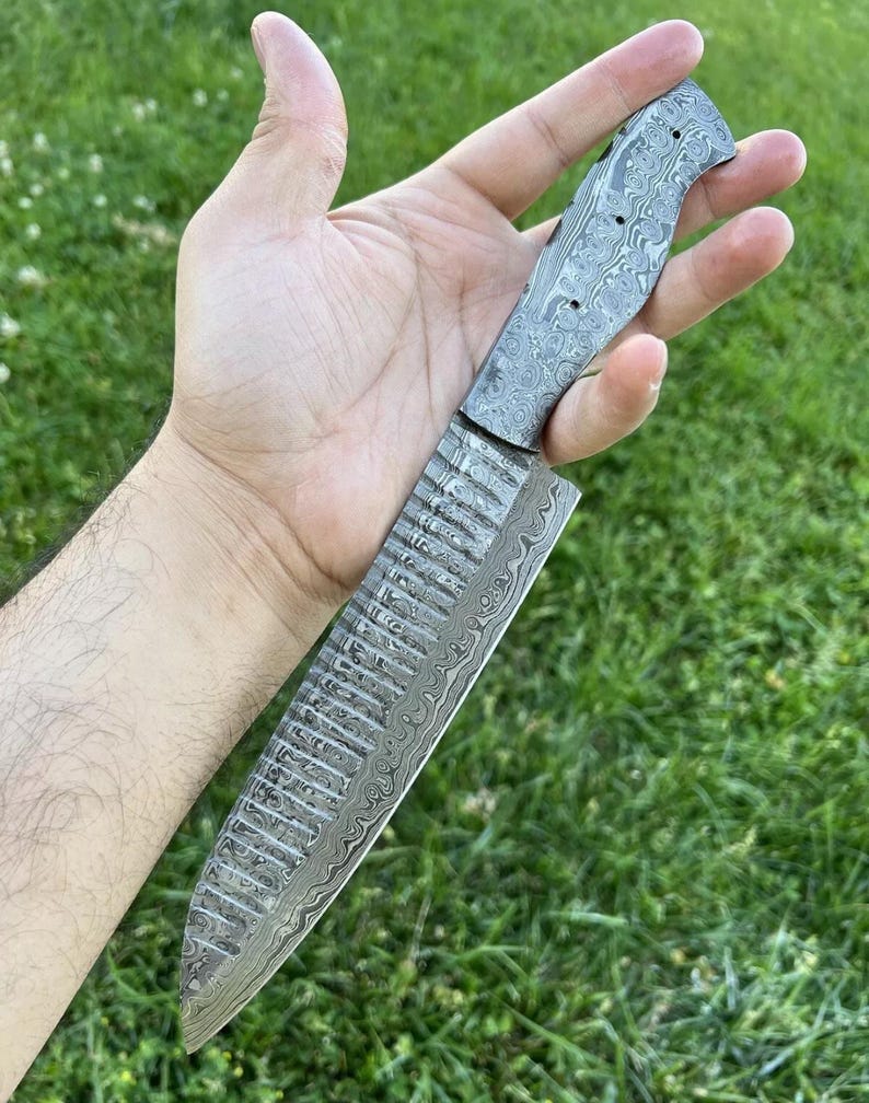 May include: A close-up of a silver knife with a patterned blade and a textured handle. The knife is held in a hand against a green grassy background.