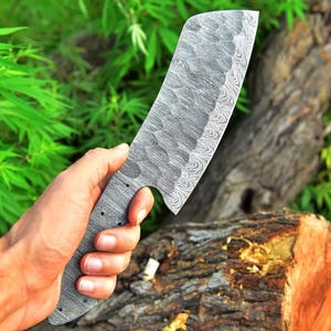 May include: A large, silver,  damascus steel cleaver with a textured blade and a black handle. The cleaver is held in a hand against a wooden background.