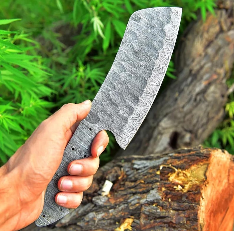 May include: A large, silver,  damascus steel cleaver with a textured blade and a black handle. The cleaver is held in a hand against a wooden background.