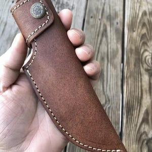May include: A brown leather knife sheath with white stitching and a metal button with a star design.