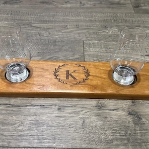 May include: A wooden tray with four glass cups and a monogrammed design. The tray is made of cherry wood and has a natural finish. The cups are clear glass and have a round base. The monogram is a letter "K" surrounded by a wreath of leaves.