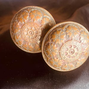 Peut inclure: Deux poignées de porte rondes en métal avec une finition bronze patiné. Chaque poignée présente un médaillon circulaire central entouré d'un motif répétitif de fleurs en relief. La surface montre des signes d'usure et d'oxydation.