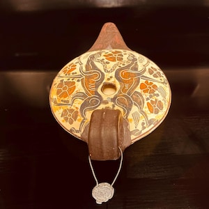May include: A ceramic oil lamp with a dark brown handle and a central fill hole. The top surface features a cream-colored background decorated with black and orange illustrations of two symmetrical animals surrounded by stylized floral patterns.