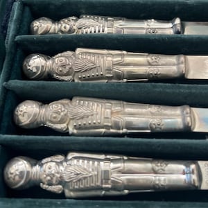 May include: Set of four silver-toned butter knives, each shaped like a detailed soldier figure. The knives are arranged in a green velvet-lined box, showcasing their intricate design and craftsmanship. The soldier figures have helmets and uniform details.