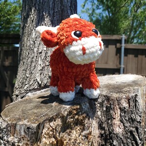 May include: A crocheted plush toy in the shape of a bull. The bull is orange with white accents and black eyes. The toy is sitting on a tree stump.