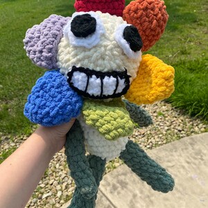 May include: A handmade, colorful crochet flower plushie with a smiling face. The flower has petals in red, orange, yellow, green, blue, and purple. The face is cream-colored with black eyes and a white-toothed grin. The stem and legs are green.