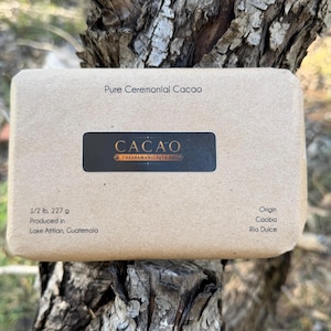 May include: A rectangular block of Pure Ceremonial Cacao wrapped in light brown paper. A black label features the text CACAO and THESHAMANICPATH.ORG. Text indicates 1/2 lb, 227 g, produced in Lake Atitlan, Guatemala, with origin Cooba, Rio Dulce.