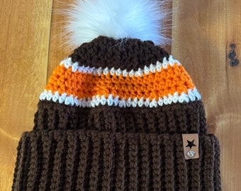 May include: Brown crocheted beanie with white pom pom and orange and white stripes. The beanie has a brown leather patch with a white star on it.