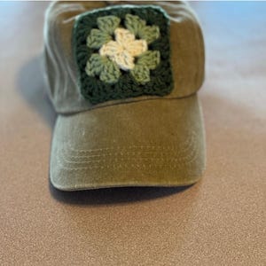 May include: Olive green baseball cap with a crocheted square patch. The patch features a floral design in shades of green and cream. The cap has a curved brim and a classic, casual style.