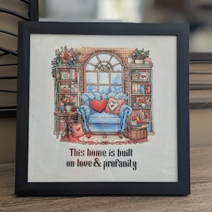 May include: A framed cross-stitch artwork featuring a cozy living room scene. The image includes a blue armchair with heart-shaped pillows, bookshelves, and a window. The text at the bottom reads, "This home is built on love & profanity."