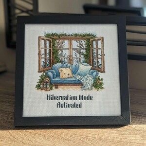 Puede incluir: Imagen de punto de cruz enmarcada en negro. Muestra un sofá azul con cojines y una manta frente a una ventana. El texto "Hibernation Mode Activated" está debajo. La ventana abierta revela un paisaje invernal.