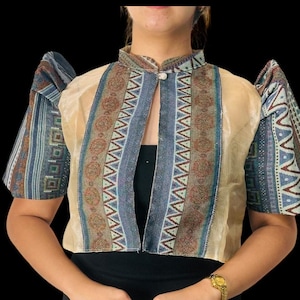 Può includere: Top beige a maniche corte con colletto alto e dettagli fantasia blu, marrone e bianchi. Il top ha una chiusura con bottoni e maniche a sbuffo. L'indumento è indossato sopra un top e una gonna neri.