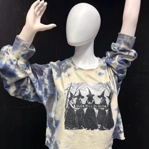 Puede incluir: Una sudadera teñida con tinte anudado en azul y crema. La camisa presenta un gráfico de cinco mujeres con sombreros de bruja con el texto "Girls Will Be Girls."