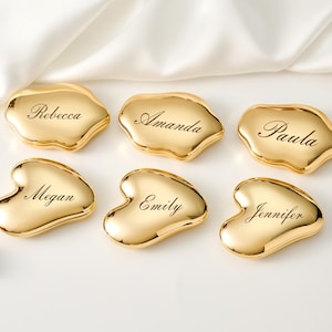 May include: Six gold-colored place card holders in organic shapes, each engraved with a different name: Rebecca, Amanda, Paula, Megan, Emily, and Jennifer. The holders are arranged on a white surface with a satin fabric.