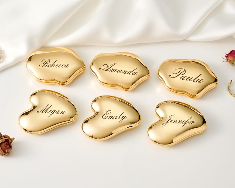 May include: Six gold-colored place card holders in organic shapes, each engraved with a different name: Rebecca, Amanda, Paula, Megan, Emily, and Jennifer. The holders are arranged on a white surface with a satin fabric.
